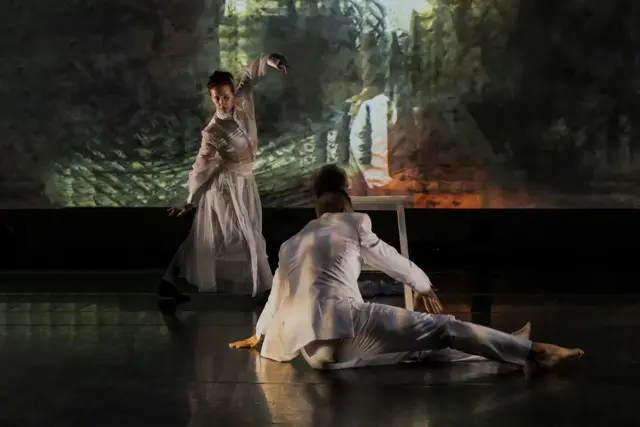 Curie Curie: two dancers in white, one kneeling, before a projected forest of green light