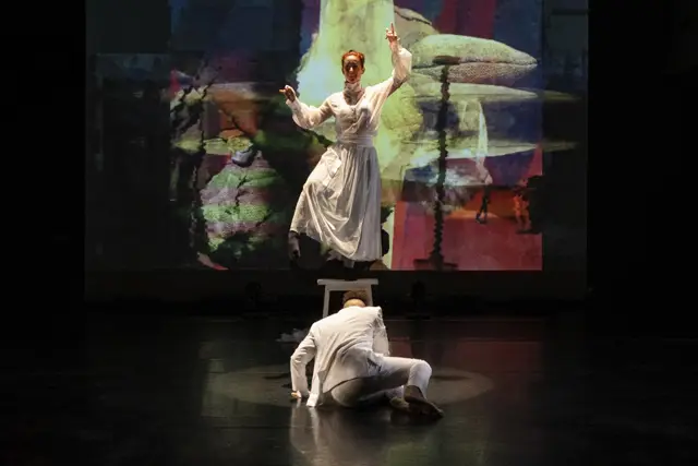 Curie Curie: a dancer with raised arms before a projected collage, a second dancer crouched below