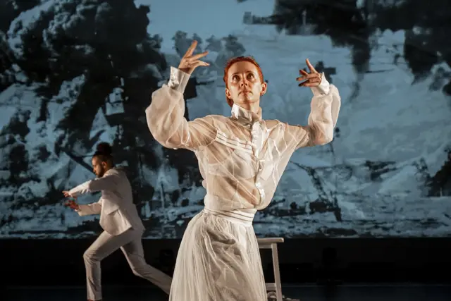 Curie Curie: a dancer with hands raised before a projected landscape in blue and gray