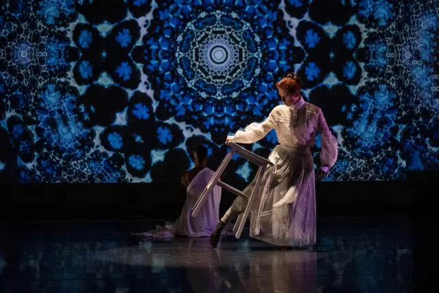 Curie Curie: a dancer turning over a stool before a blue kaleidoscopic projection