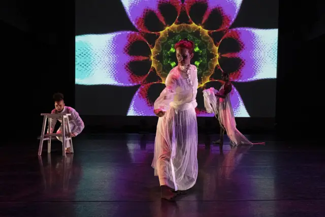 Curie Curie: a dancer in white before a projected magenta and green flower, another seated on a stool