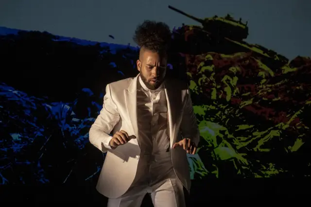 Curie Curie: a dancer in a white suit before a projected battlefield with a tank