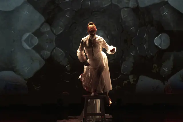 Curie Curie: a dancer standing on a stool before a projected gray stone pattern