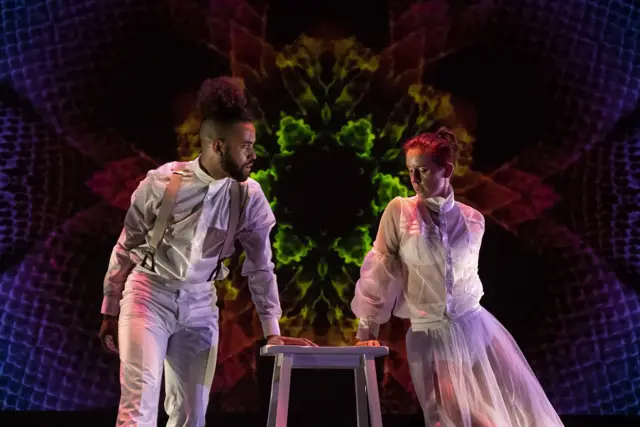 Curie Curie: two dancers at a stool before a red, green, and violet kaleidoscope